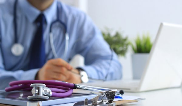 Doctor sitting down working on a laptop with a stethoscope on the table.