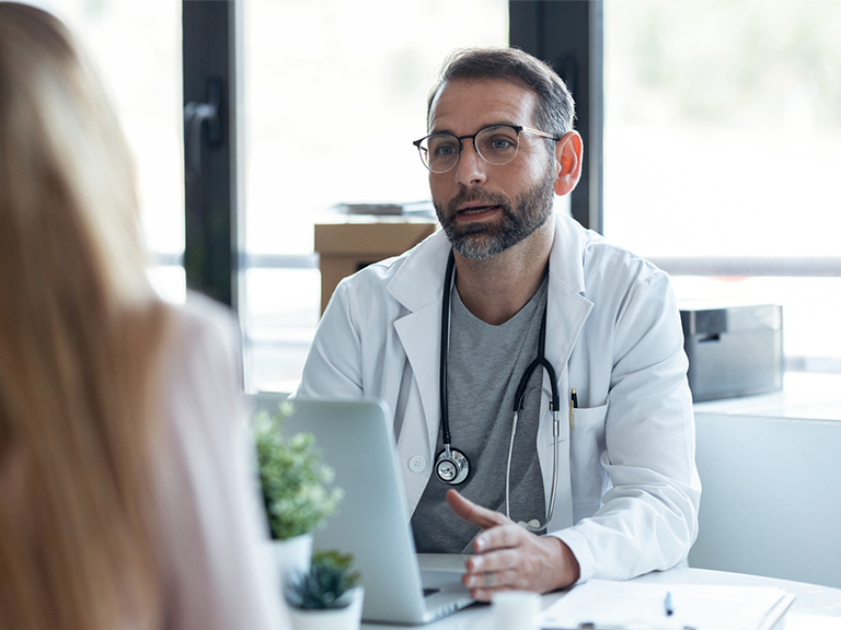 A doctor sitting behind a desk with a laptop, explaining something to a patient on the other side.